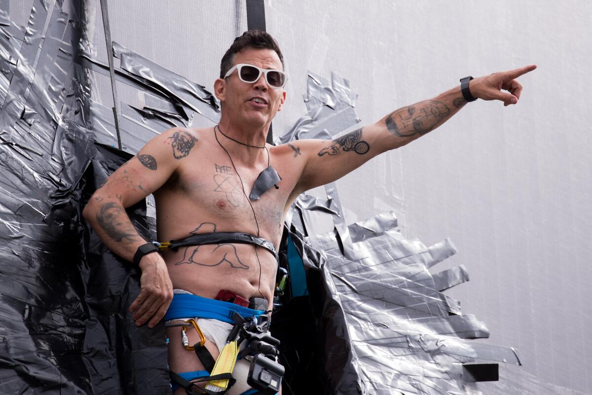 Steve-O, shirtless and wearing white-rimmed sunglasses, points while duct-taped to a structure
