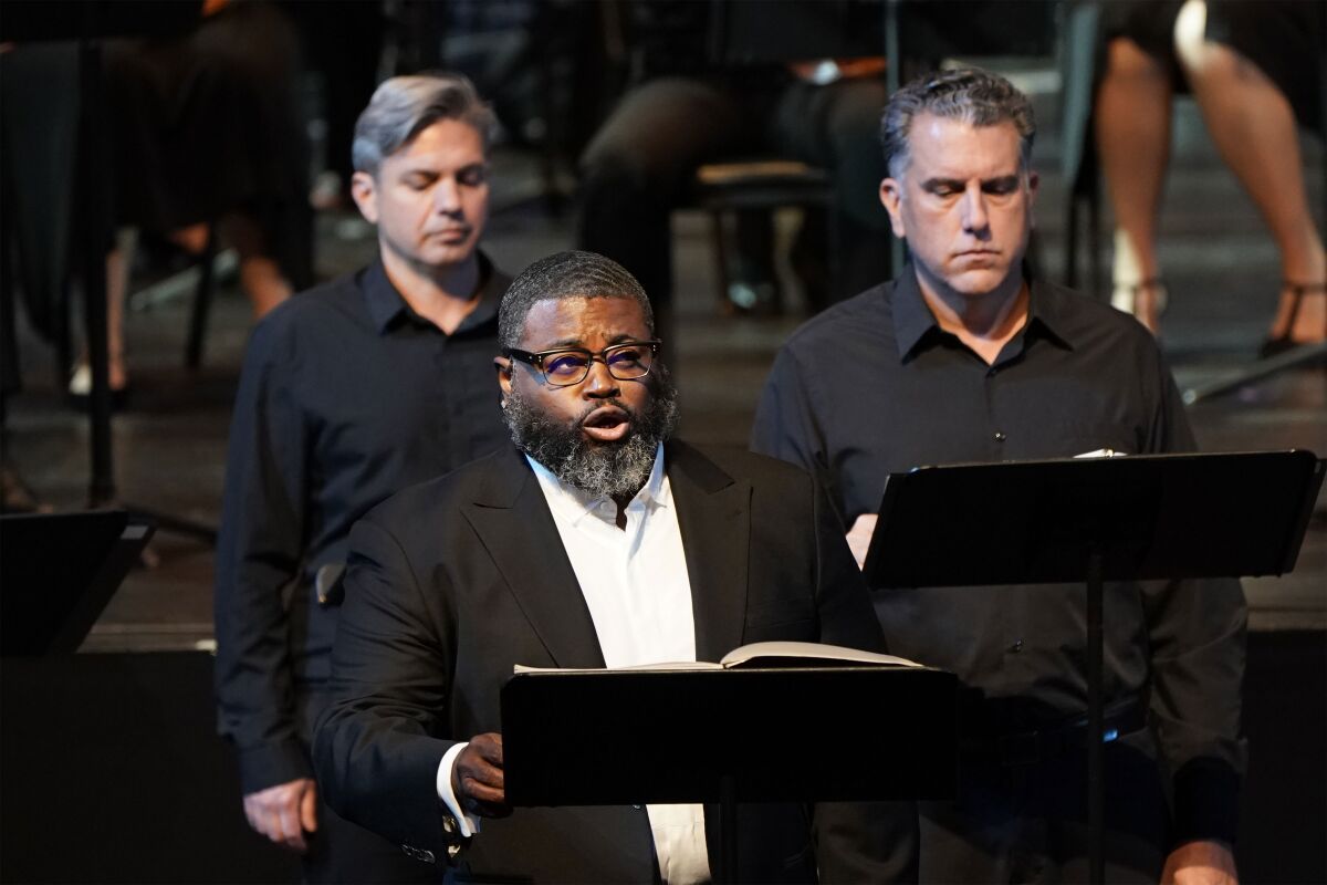 Russell Thomas, dressed in a black suit, sings before a music stand as other performers flank him