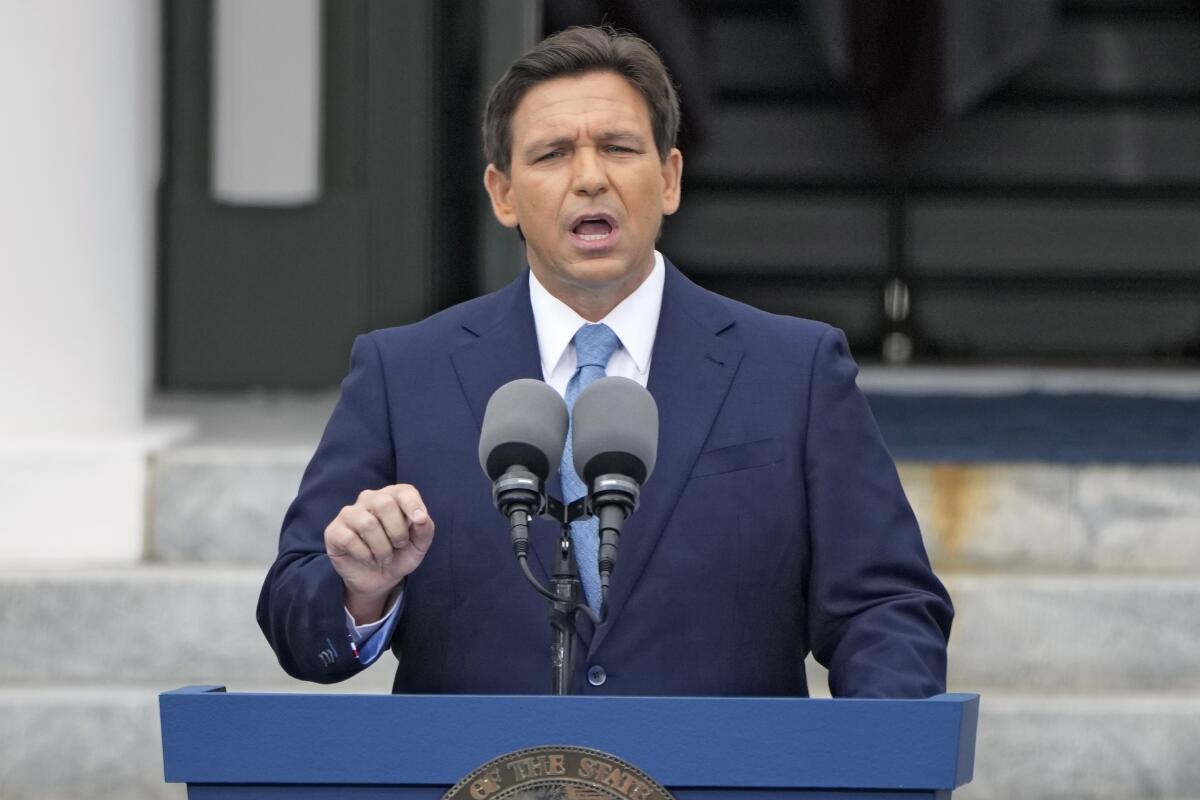 A man with dark hair, in a navy blue suit and light blue tie, speaks at a lectern 