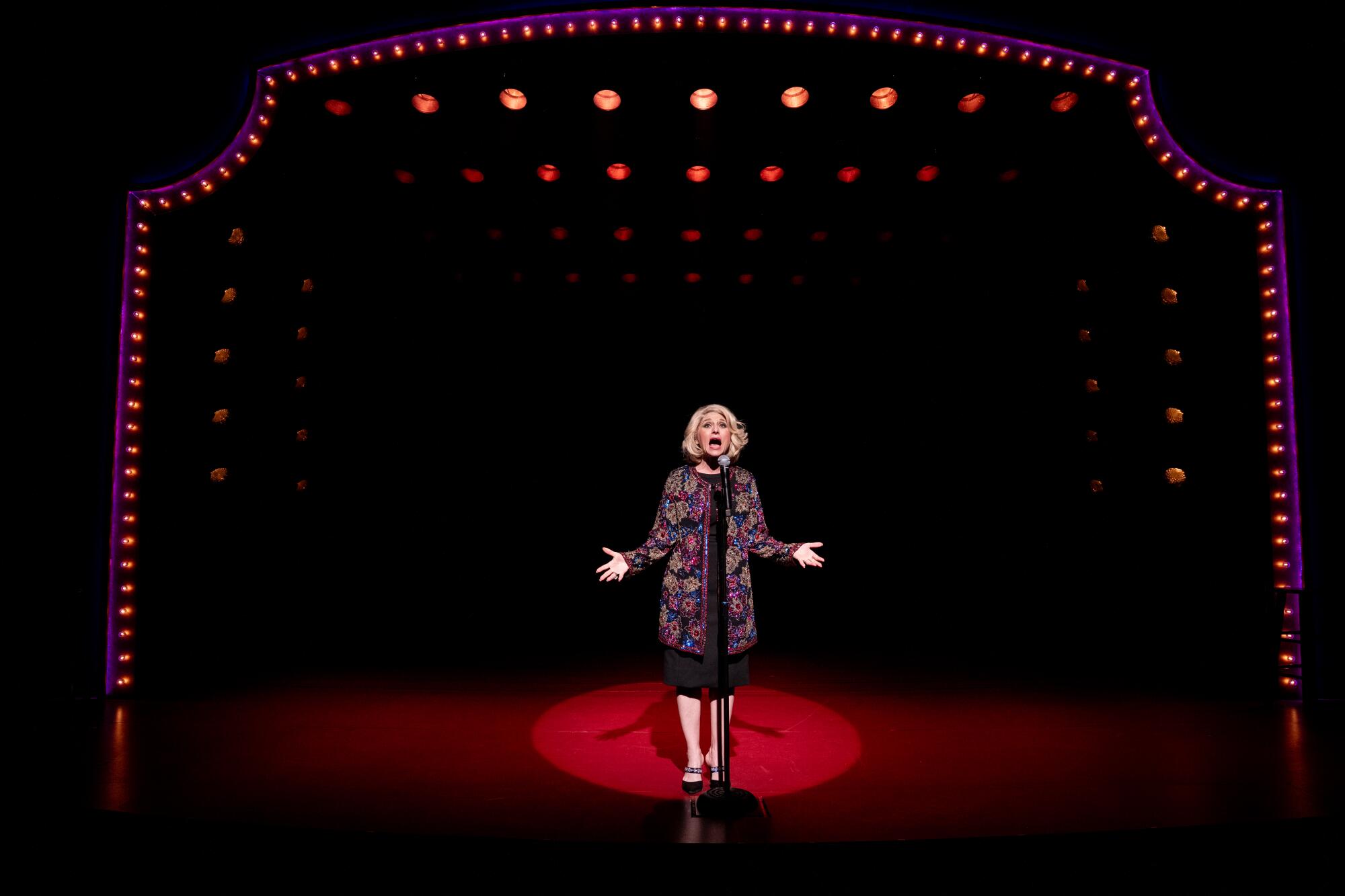 A blond woman stands onstage talking into a stand microphone, under a proscenium arch outlined in lightbulbs