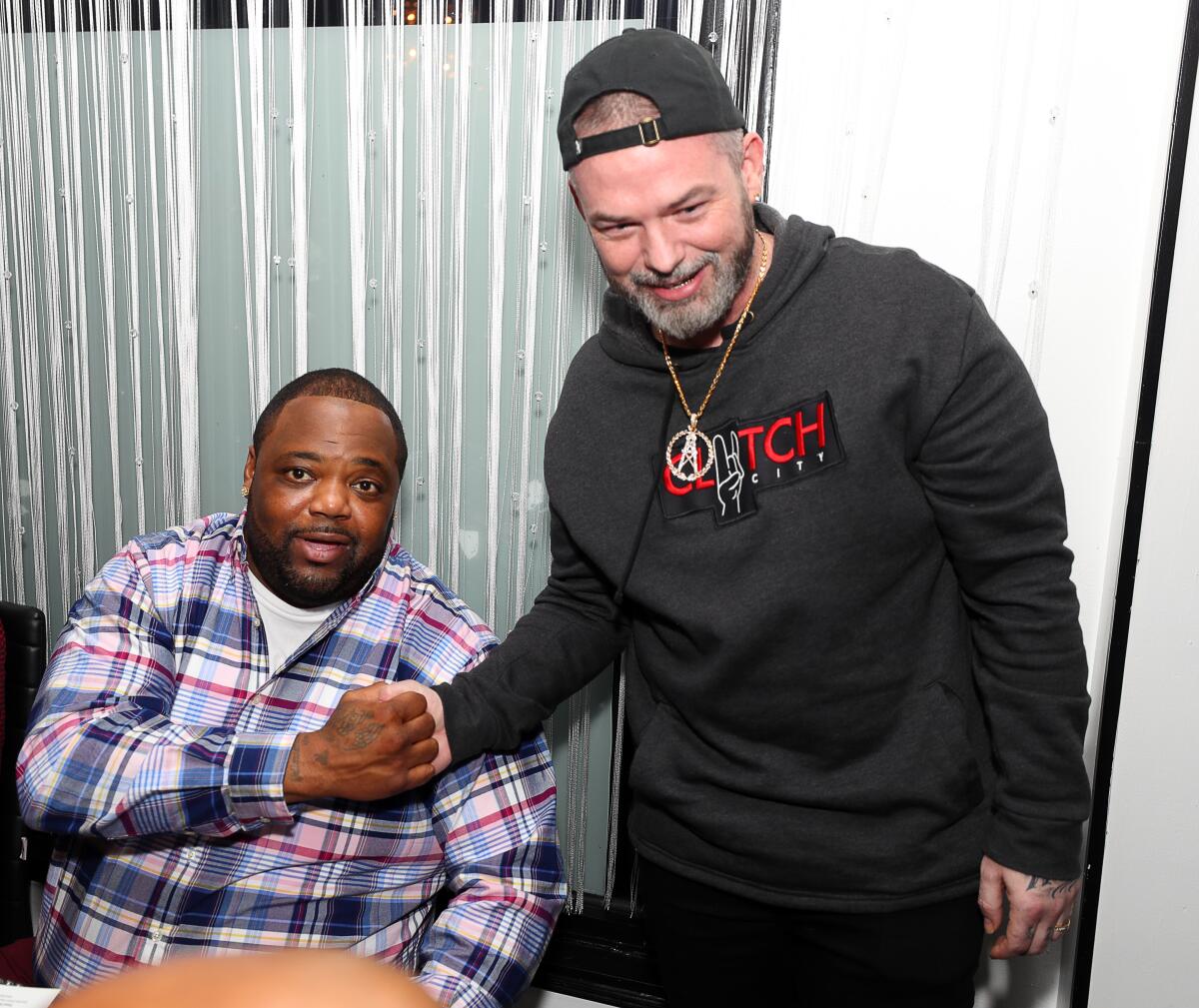Big Pokey sits and shakes hands with Paul Wall, who is standing.