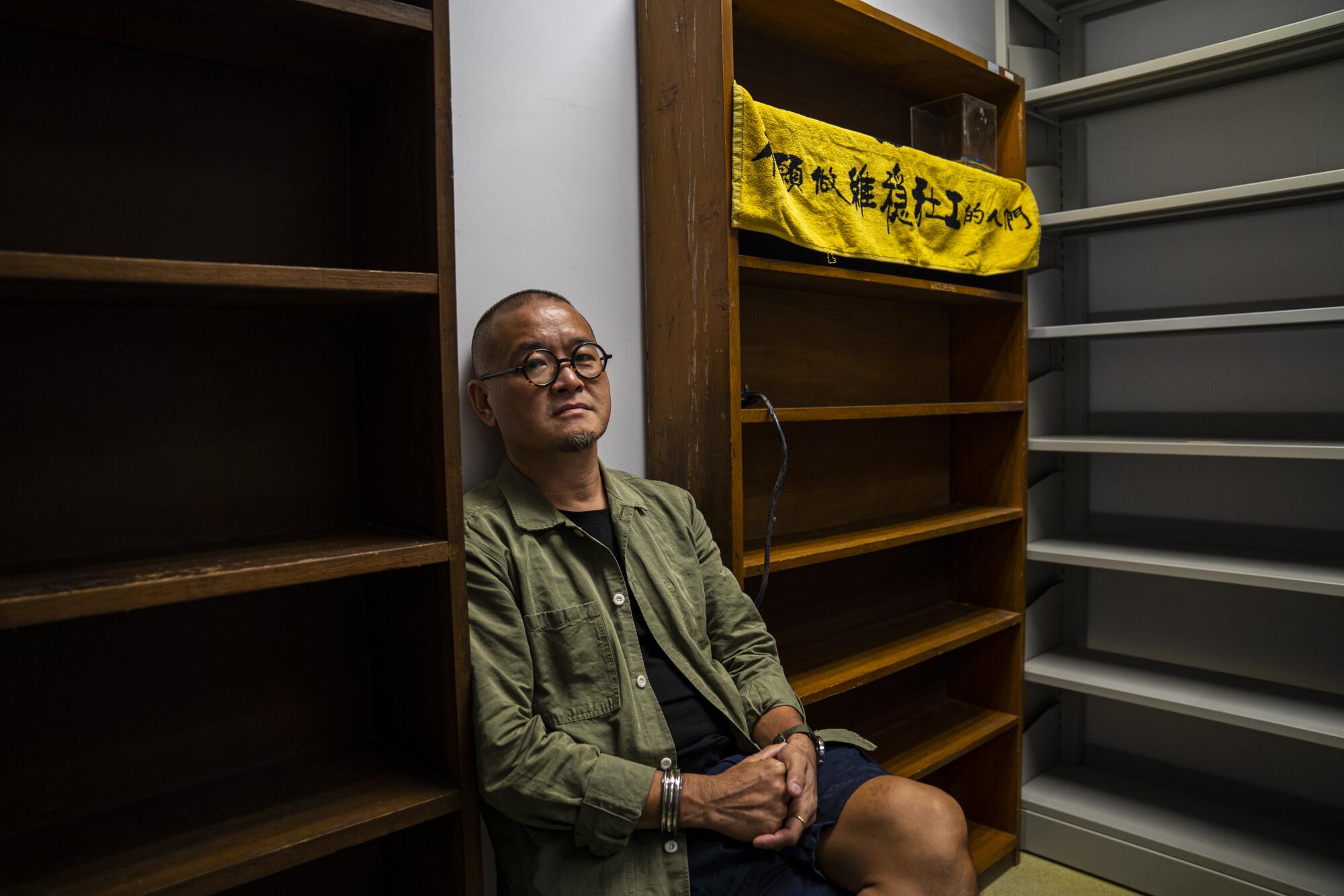 A man in glasses and a green overshirt sits in between empty bookshelves.