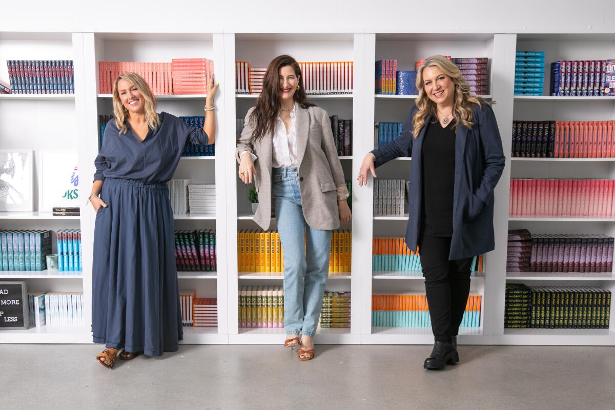 Three women (Liz Tigelaar, Kathryn Hahn, Cheryl Strayed) pose in front of colorful books, separated by the shelves.
