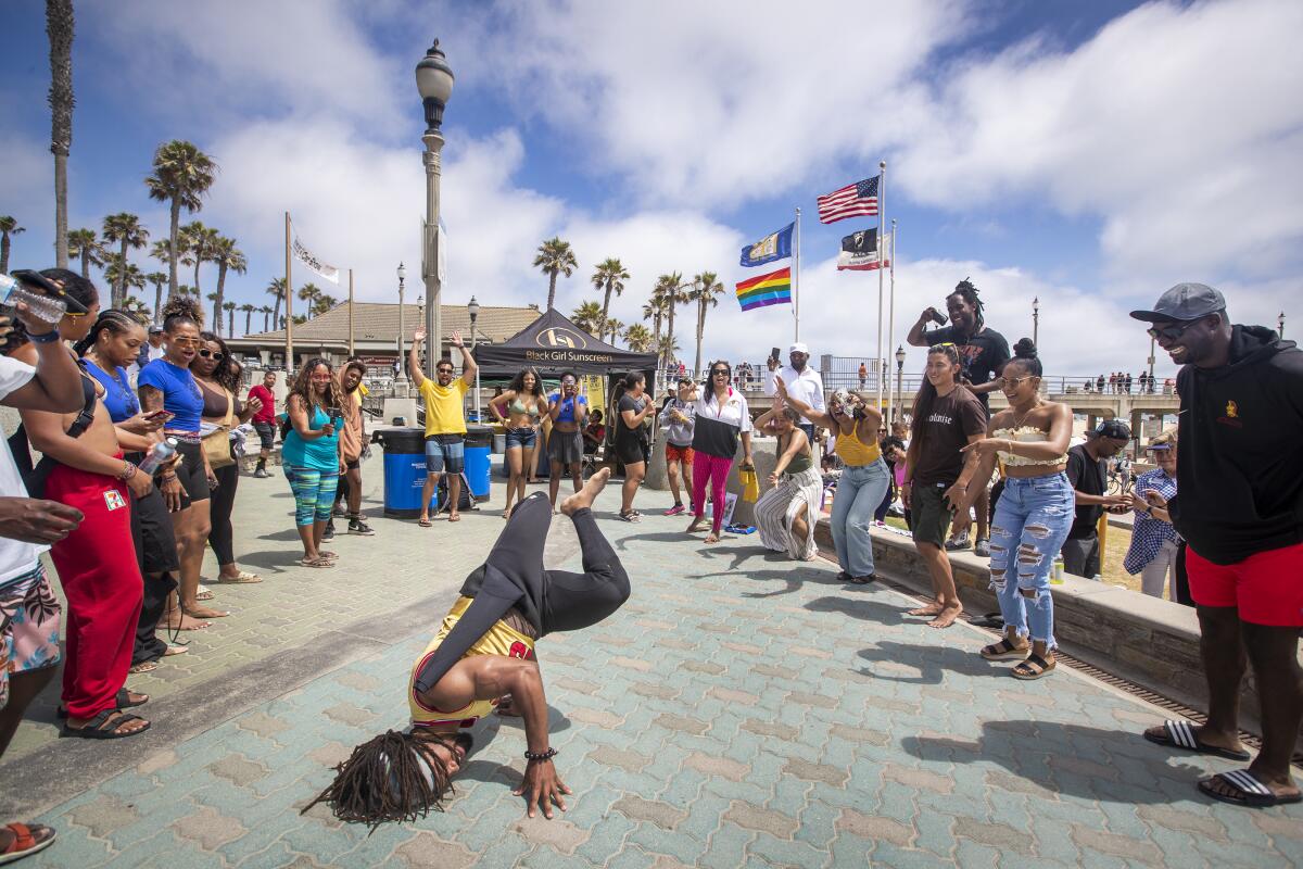 A man in a wetsuit performs a breakdance move on a sidewalk by the beach as a crowd of people cheer him on.