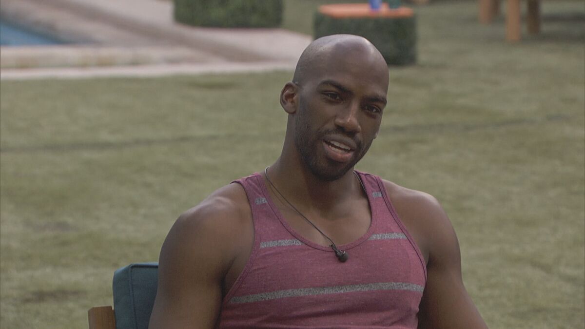 A man in a maroon tank top sitting outside with Astroturf behind him