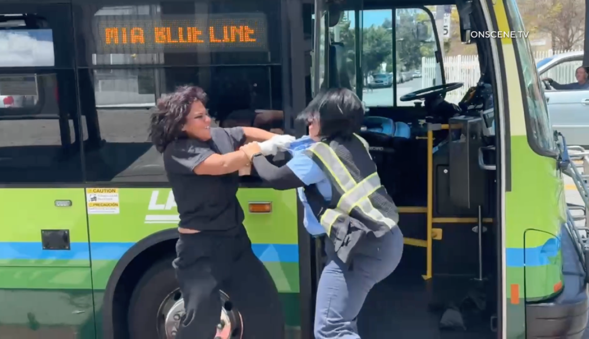 Two women, one in a reflective vest, grasp arms in a fight outside a green bus.