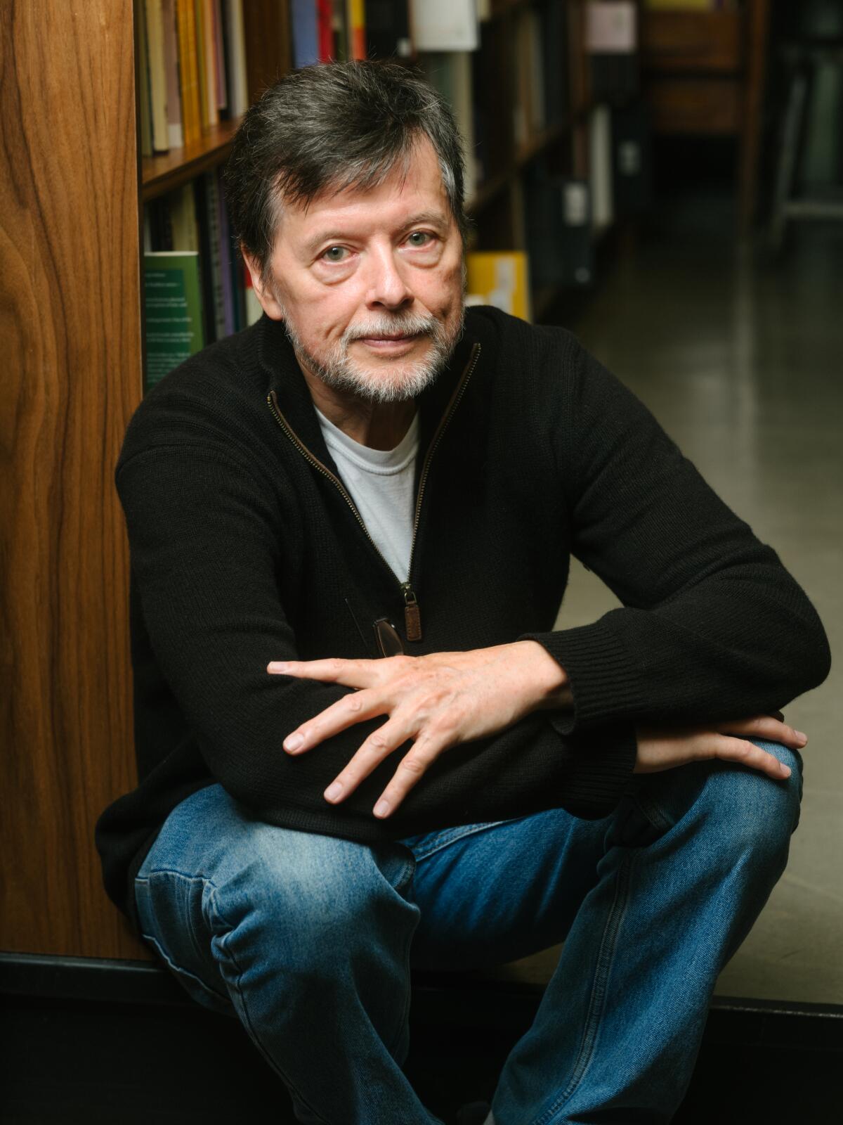 Documentarian Ken Burns in a black sweater and blue jeans, crouched by a shelf of books in a bookstore.