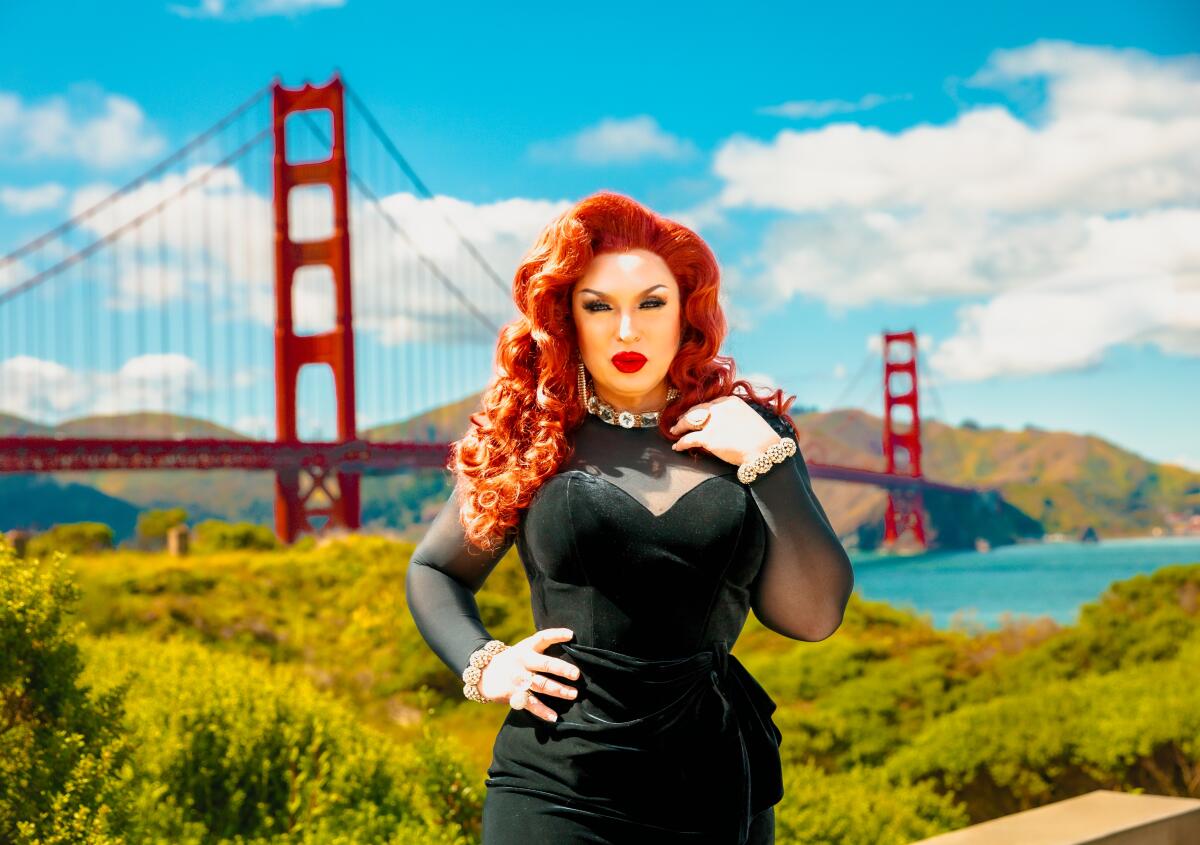 A person with long red hair, wearing a velvet black dress, poses for a photo in front of the Golden Gate Bridge.