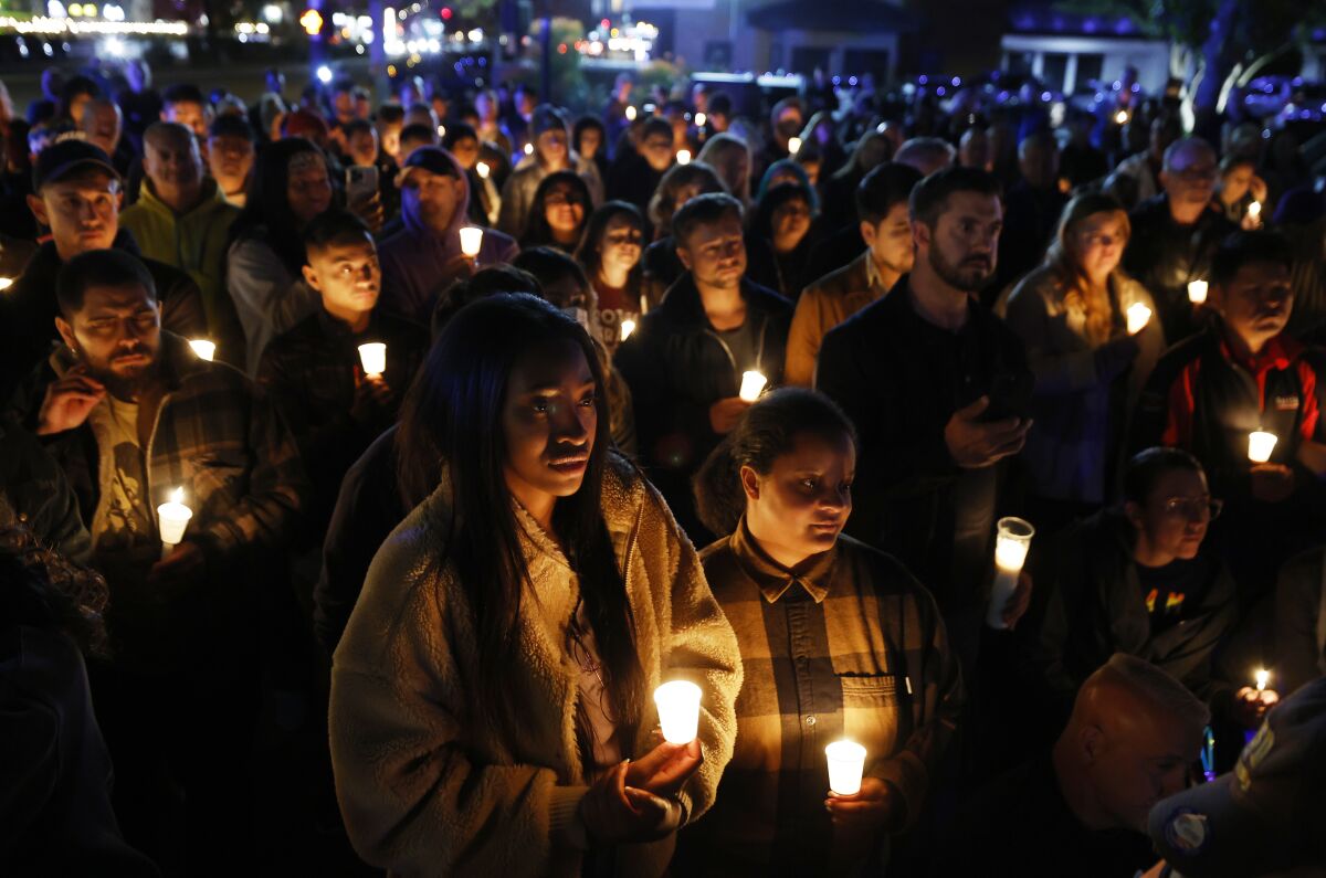 A crowd of people, their faces somber, gather outside, holding candles.