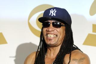 Melle Mel is posing, flexing his arms while wearing a black tank top, silver chains, a Yankees cap and dark sunglasses