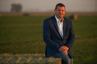 A man in a jacket and shirt, hands clasped, smiles while sitting on the edge of a bale of hay
