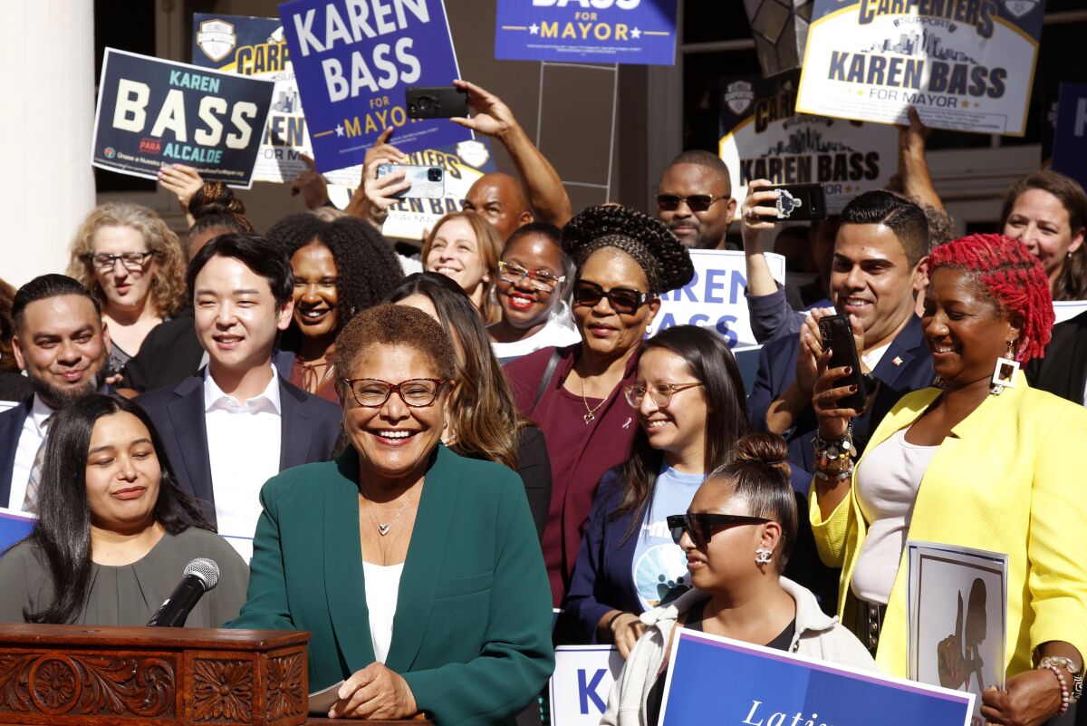Karen Bass smiling and standing by a microphone, with a crowd of supporters behind her, many holding campaign signs
