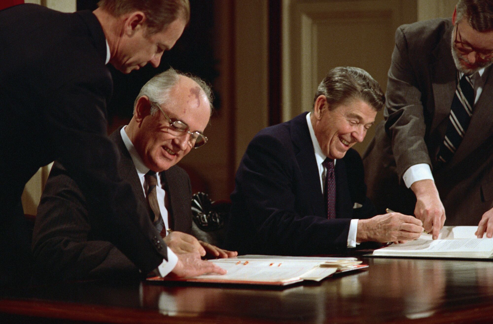 One man in suit and tie and glasses, left, and another man, also in suit and tie, smile as they prepare to sign documents 