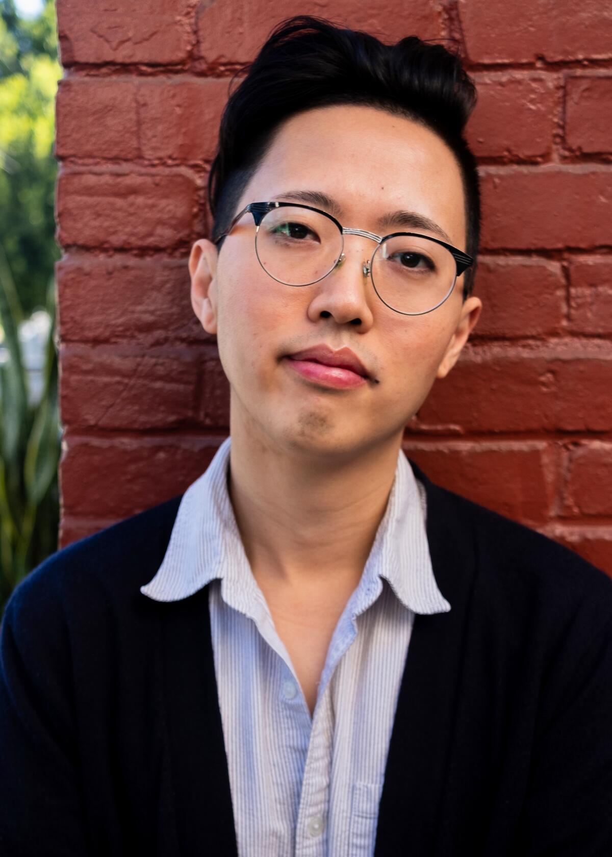 A headshot of a person with glasses leaning against a brick wall.
