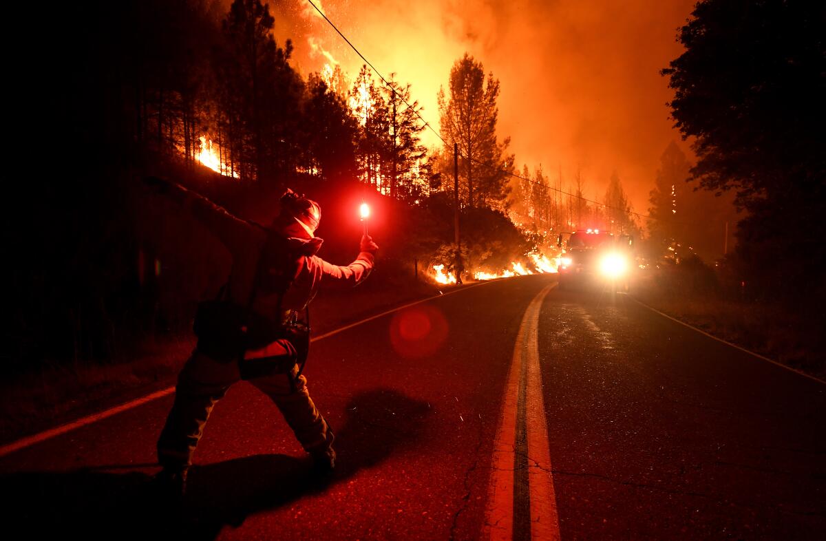 A firefighter cocks his arm back to throw a lit flare as massive orange flames burn along a two-lane highway in the woods