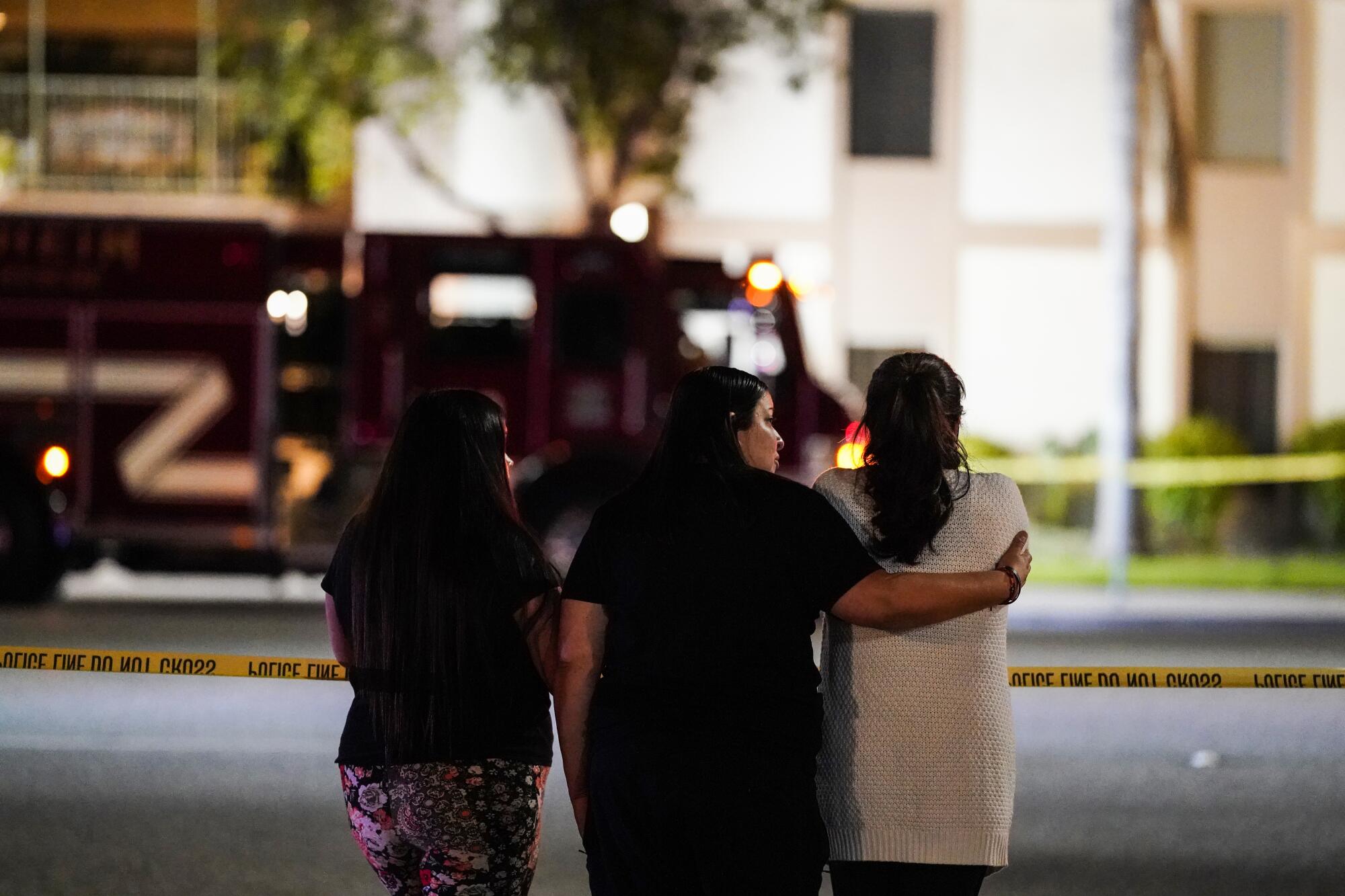Three women, one with her arm around another, stand near the scene of the shooting.