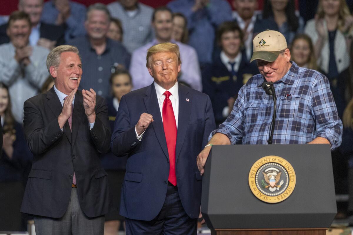Kevin McCarthy and Donald Trump stand next to a man in a plaid shirt and baseball shirt at a podium.