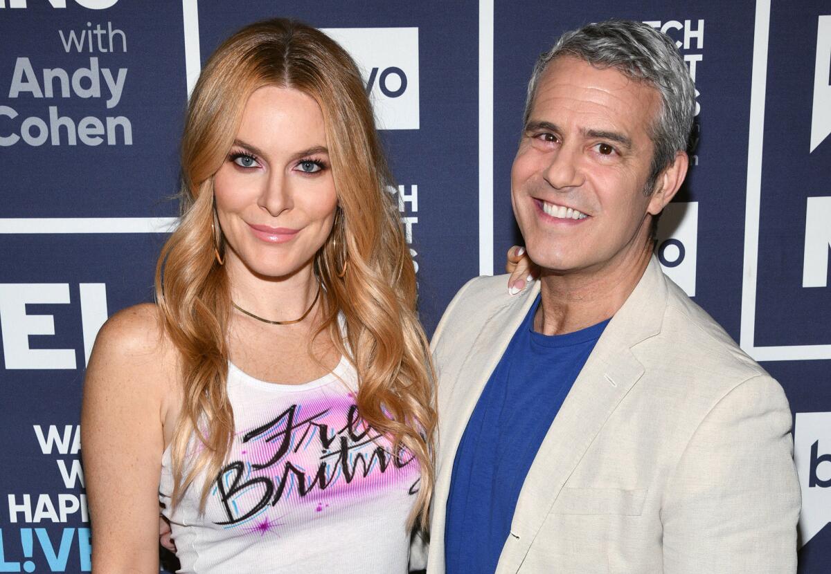 A woman with wavy blond hair in a white tank top standing next to Andy Cohen in a beige suit jacket and blue shirt
