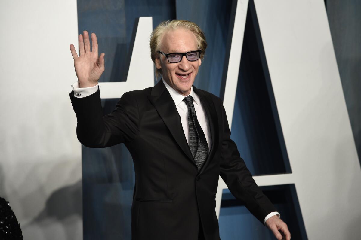 Bill Maher waves and smiles as he walks by in a black suit and tie with a white collared shirt