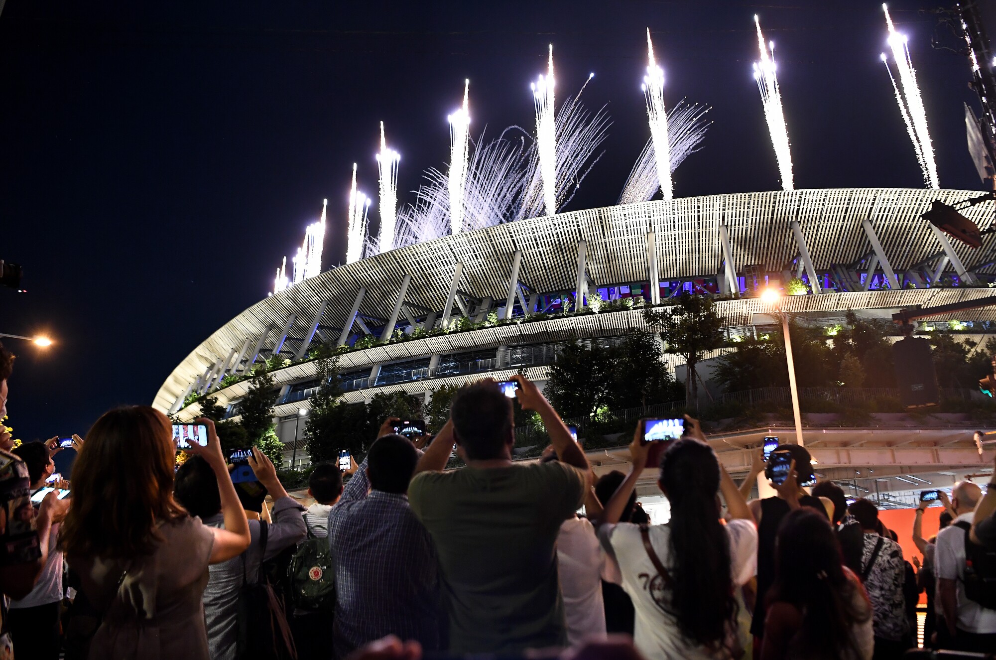 A crowd of people outside a stadium hold up their phones as fireworks explode overhead.