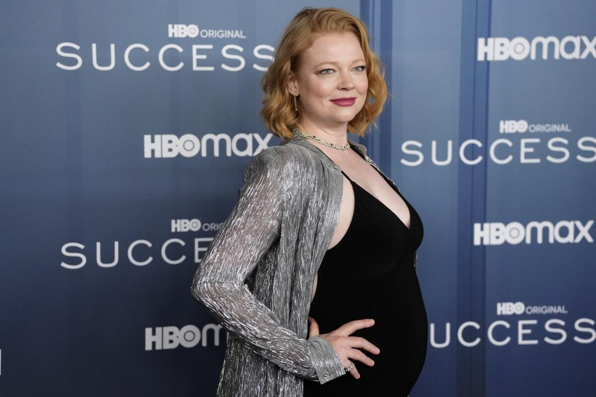 A pregnant woman with short red hair posing with her hands on her hips in a tight black dress and silver shrug
