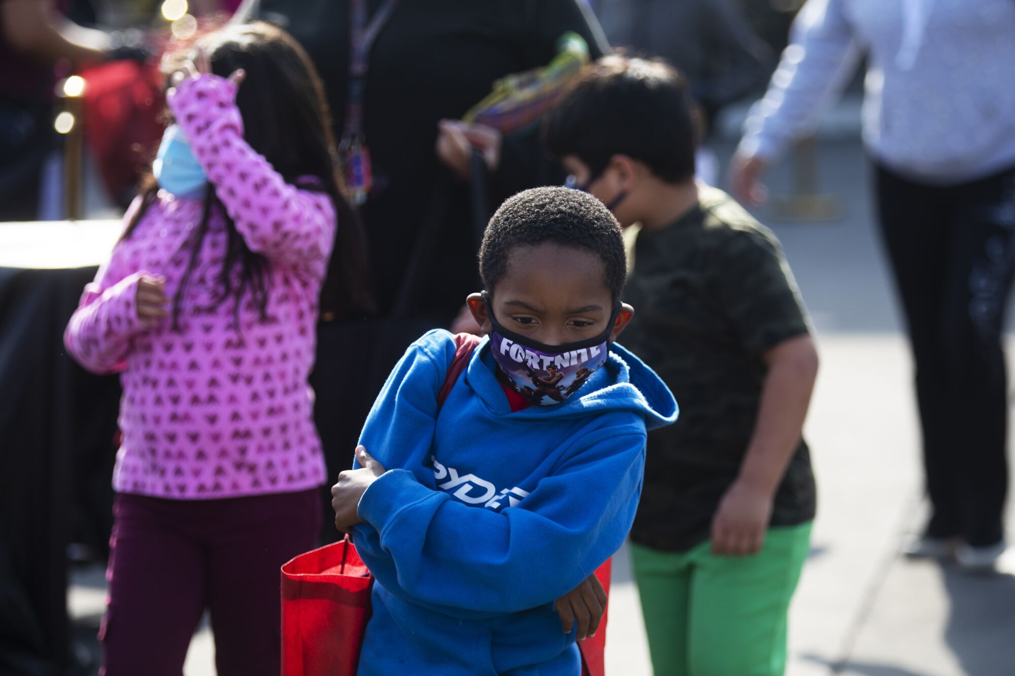 A little boy in a blue sweatshirt and Fortnite masks crosses his arms with a red tote bag over his shoulder