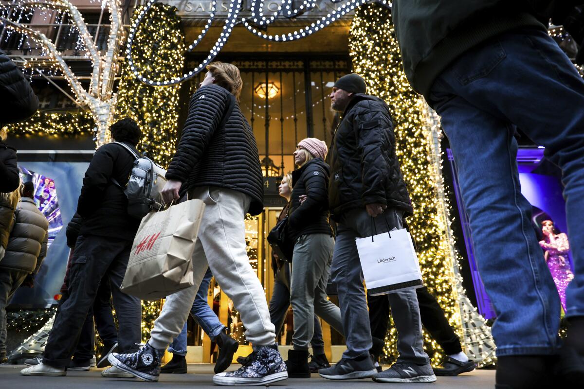Shoppers carrying bags of merchandise along a sidewalk past stores and trees decorated in lights