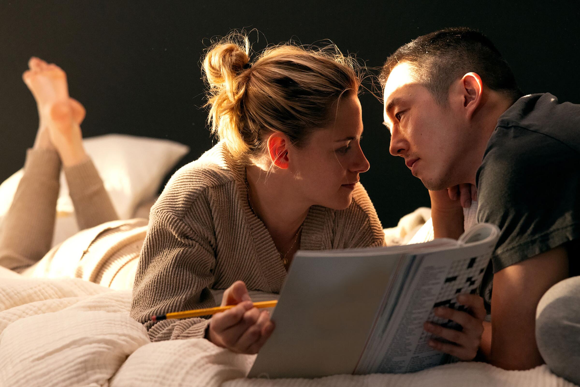 A woman lies on a bed reading a magazine, with a man next to her looking into her face.