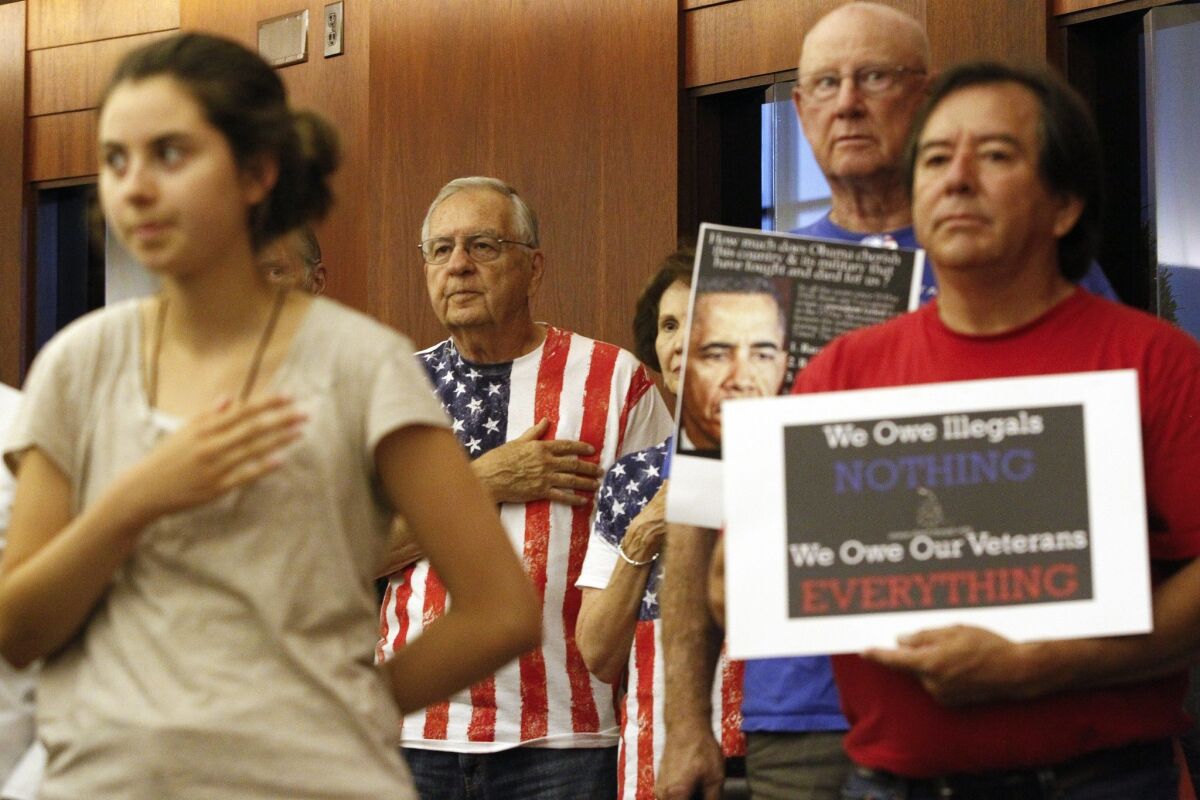 Those against the shelter for immigrant children, back and at right, hold their hands over their hearts during the Pledge of Allegiance before the start of the Escondido Planning Commission meeting.