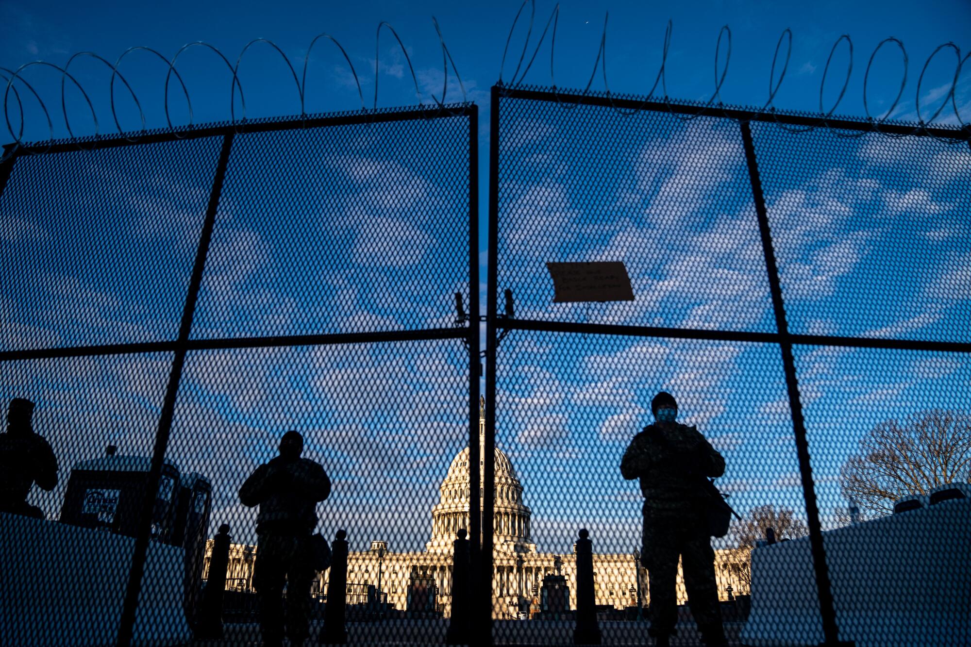 ational Guard troops stand behind security fencing with the dome of the U.S. Capitol Building behind them.