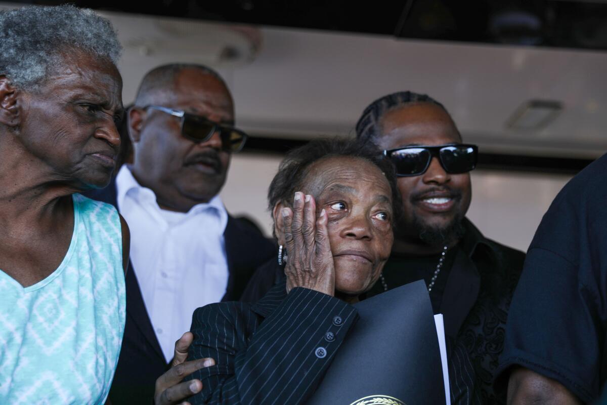 A woman stands with a hand on her cheek as she is pulled close by a man standing next to her among a group of people