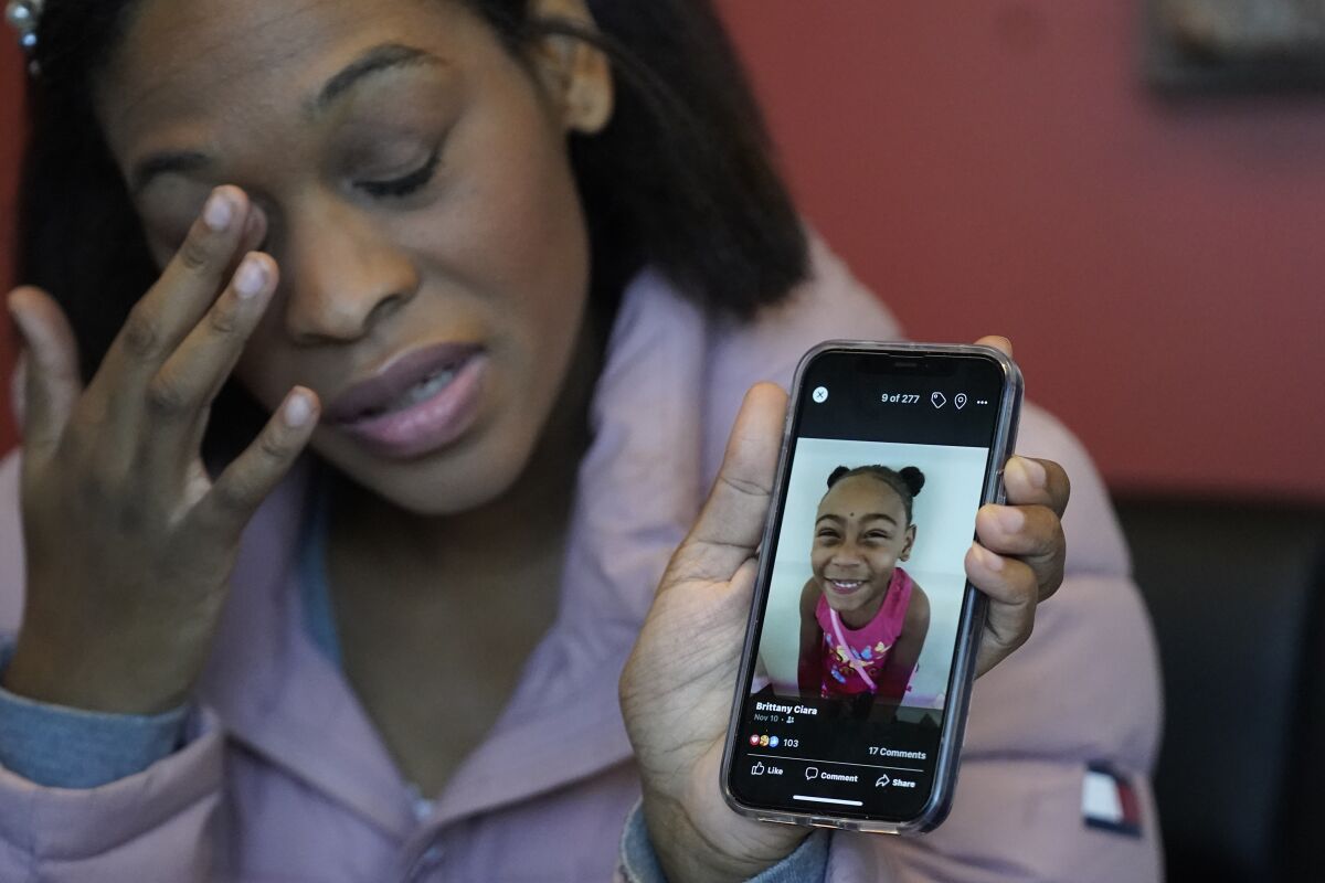 A woman wipes her eye as she holds up a cellphone showing a picture of a smiling girl in a pink top