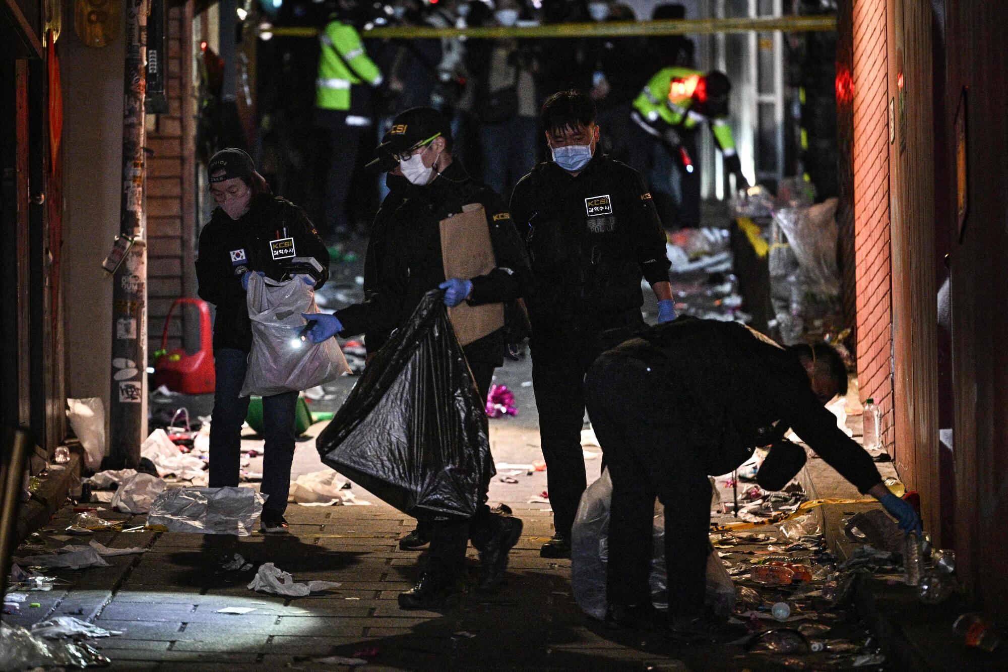 Uniformed crime scene investigators carry plastic bags and collect items from the ground in a debris-filled alley 