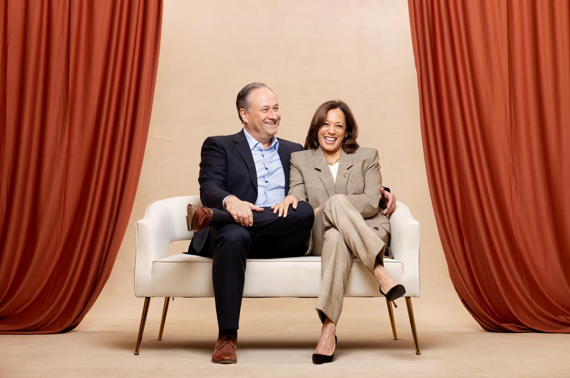  Vice President Kamala Harris and Second Gentleman Doug Emhoff sit on a couch with reddish-orange drapes on either side.