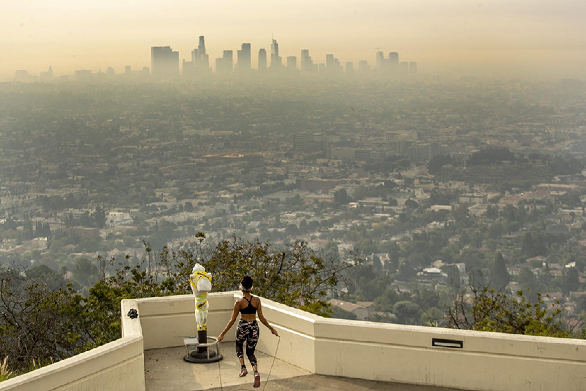 Wildfire smoke obscures a city skyline as a woman jumps rope in the foreground.