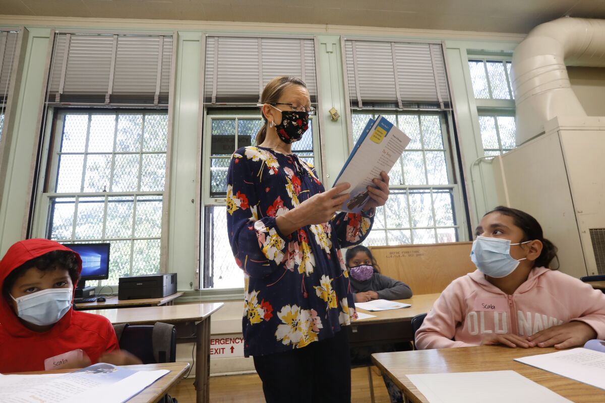 A teacher stands in class between two students. All are wearing masks.