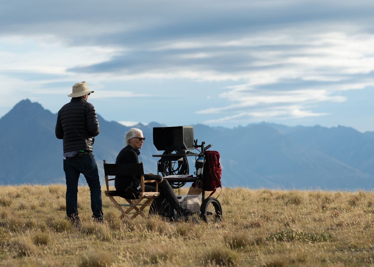 On a grassy plain with mountains beyond, a man looks over the shoulder of a woman seated behind a movie camera.