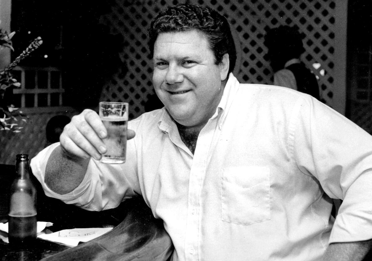 George Wendt smiles while sitting at a bar holding up a glass of beer.