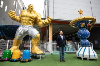 A man is flanked by a statue of a golden giant with muscles in silver shorts, left, and a sculpture of stacked blue spheres