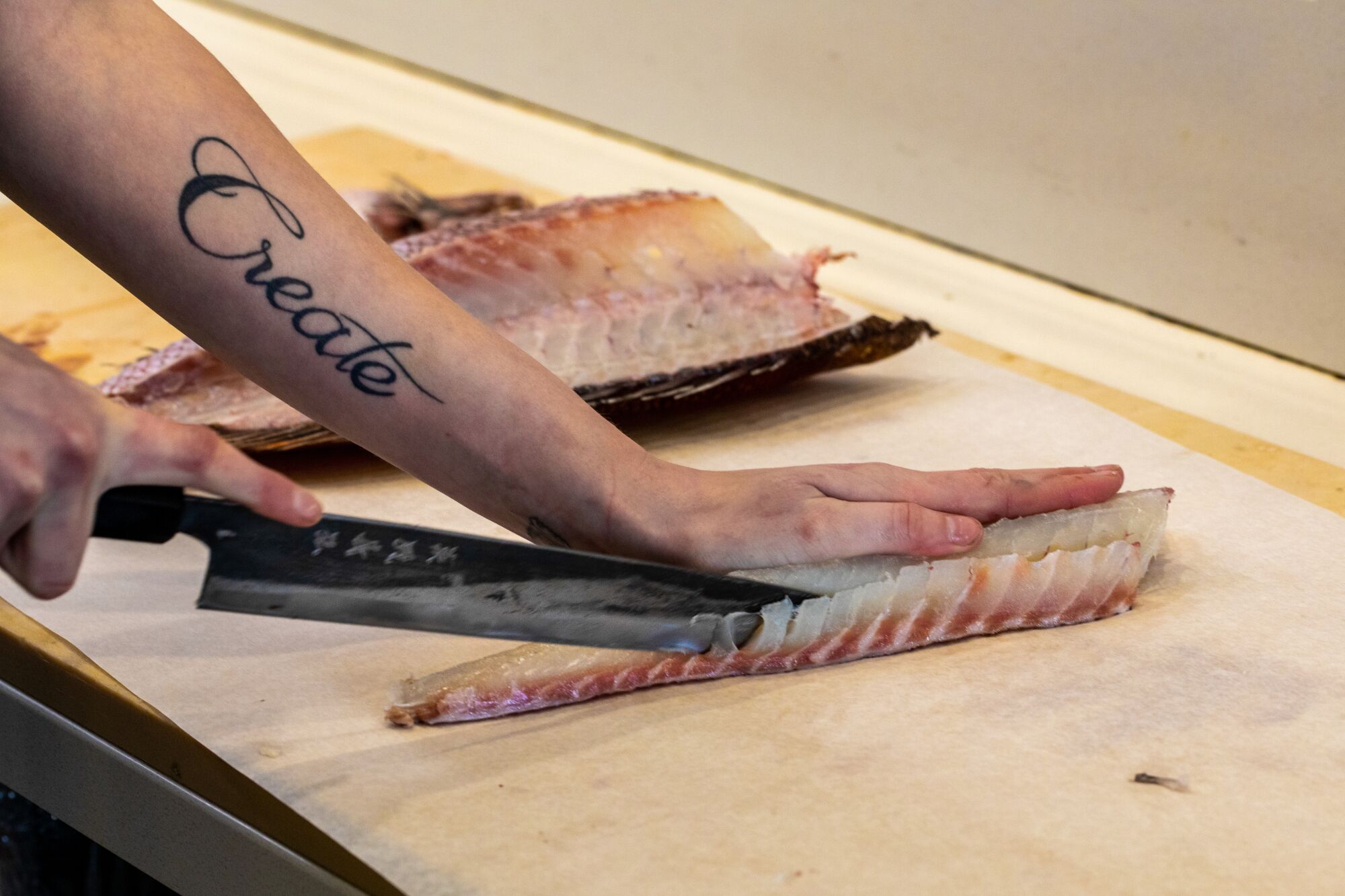 Fish is trimmed at a seafood market. The employee's arm features a tattoo saying "Create" in cursive.