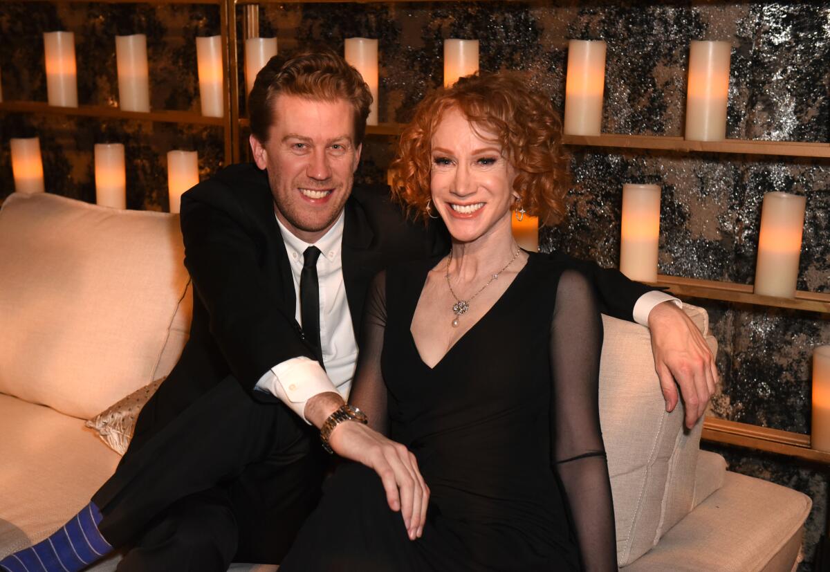 Randy Bick in black suit and tie and Kathy Griffin in a black dress with sheer sleeves smile as they sit on a peach sofa.