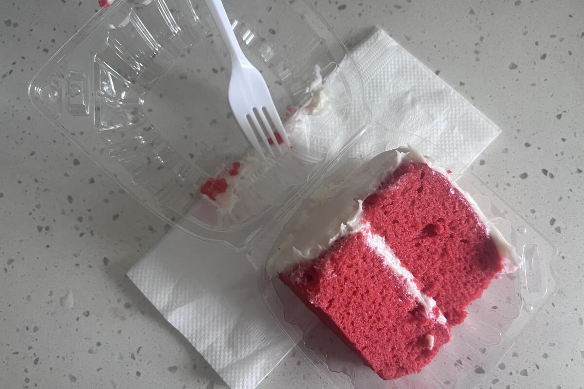 A slice of red velvet cake rests in an open clear plastic container with a white plastic fork