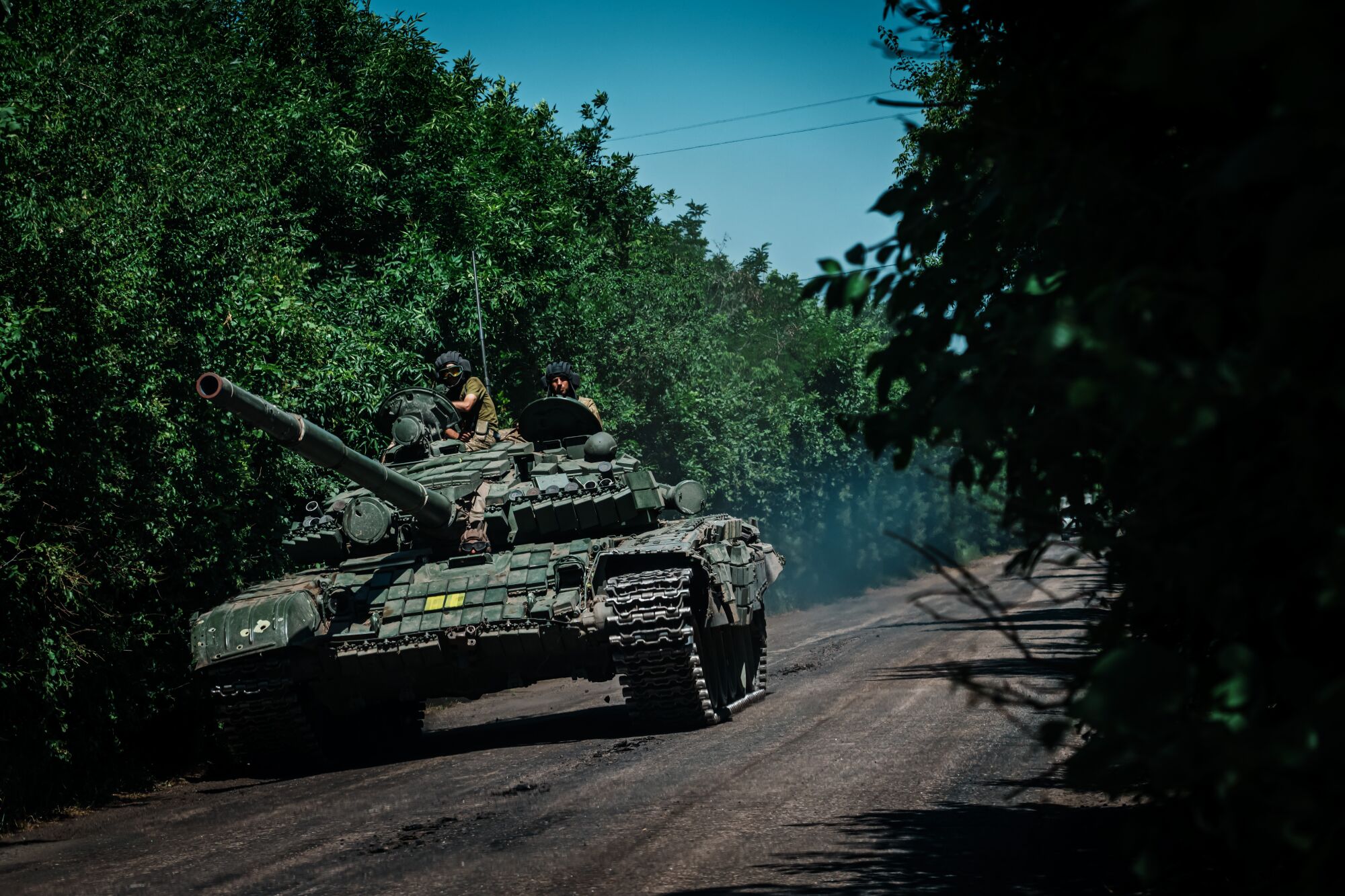 Two people in fatigues atop a tank on a road, along a dense row of green trees