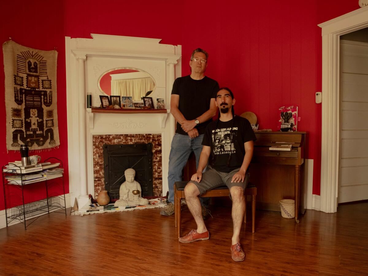 Sesshu Foster, standing, and Arturo Romo, seated, pose in a home whose walls are painted red
