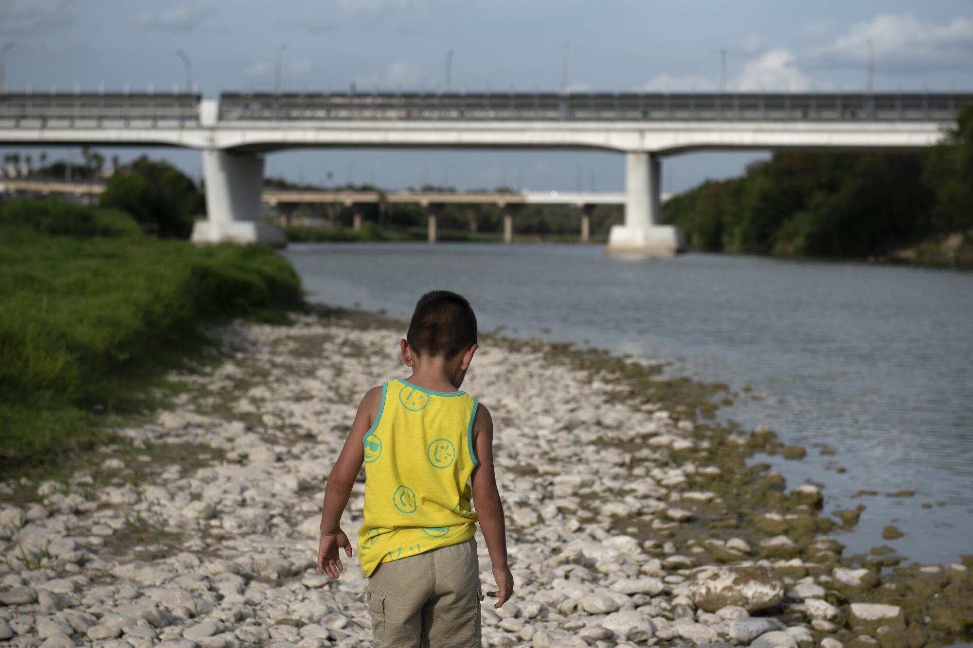 A child walks along a river approaching a bridge.