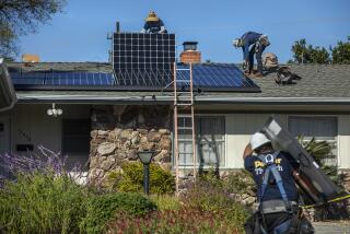 Two men install solar panels on a roof. In foreground a man carries another solar panel to be installed.
