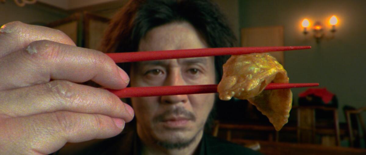 Hands use red chopsticks to hold up a piece of meat in front of a man's face