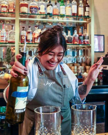 A laughing woman prepares to pour from a bottle into two glasses in front of her at a bar