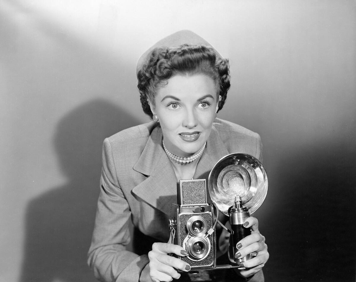 Black and white photo of a woman in a suit, pillbox hat and choker pearl necklace holding a flash camera.