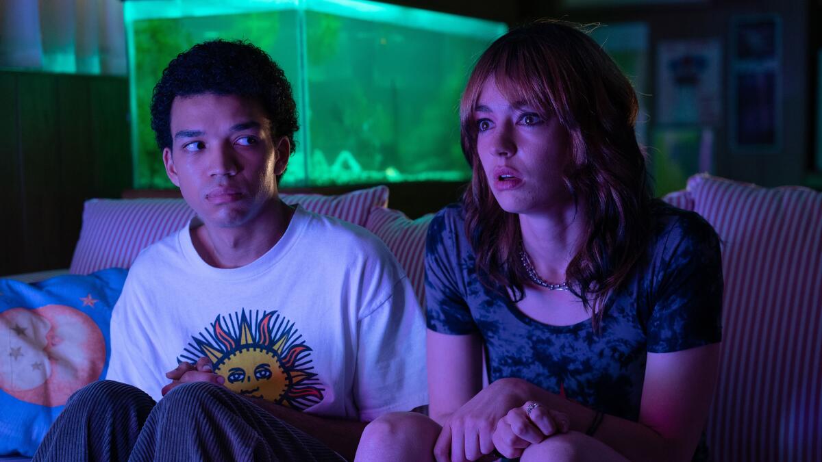 Two young people sit on a couch looking toward a TV, a glowing fish tank behind them.